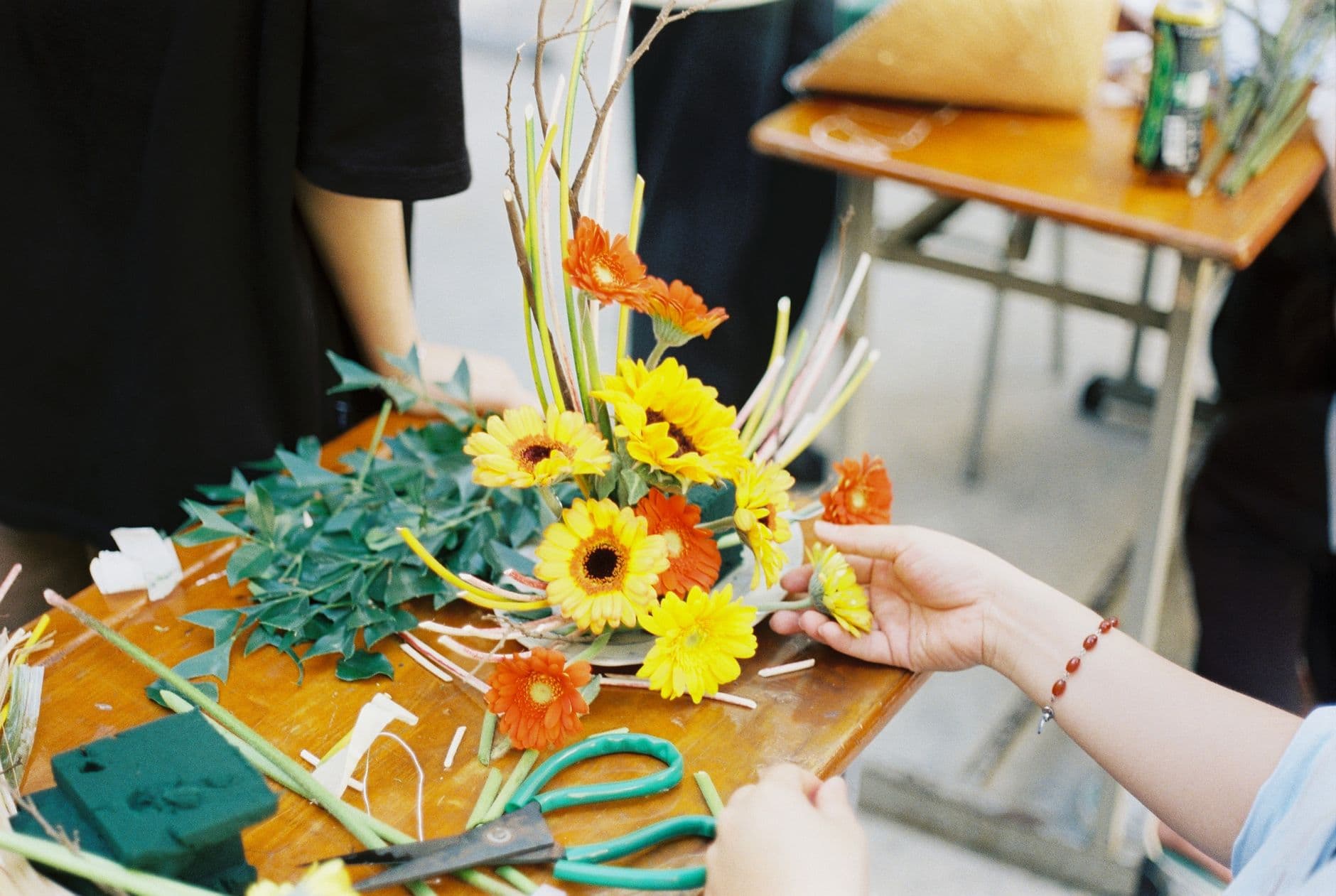 Radni stol u radionici: aranžman od gerbera i suncokreta zaboden u spužvu, uz njega bršljan, škare i odrezane stabljike.